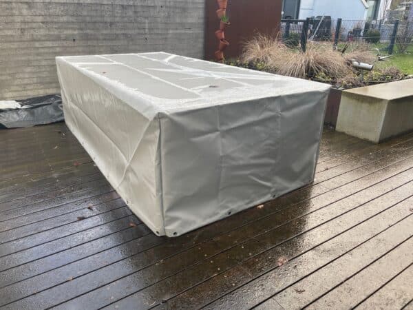 Ein rechteckiger Gartentisch mit einer beigefarbenen rechteckigen Abdeckhaube steht auf einem nassen Holzdeck. In der Nähe sorgen eine Betonbank und ein Pflanzkübel mit Gräsern für Charme, alles umgeben von einer Backsteinmauer und einem Metallzaun.