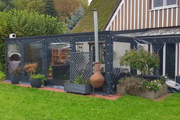 Eine gemütliche Terrasse mit transparenter Carport- oder Terrassenplane – mit 1x Fenster/ohne Tür verfügt über Hängelampen, einen Kaminofen und Topfpflanzen. Das reetgedeckte Haus liegt eingebettet zwischen Bäumen und üppigem Grün.