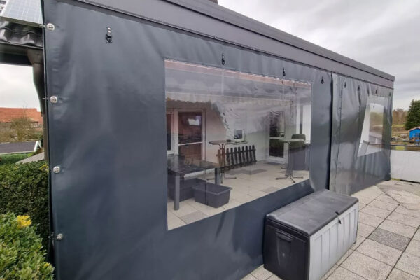Eine überdachte Terrasse mit dunklen Planenwänden und durchsichtigen Kunststofffenstern gibt den Blick auf die Möbel im Inneren frei. Die Terrassenverkleidung, oder „Carport- oder Terrassenplane – mit 1x Fenster/ohne Tür“, befindet sich auf einem gefliesten Außenbereich unter grauem Himmel.