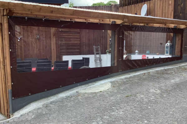 Ein Außensitzplatz mit Carport- oder Terrassenplane – mit klaren Kunststofffenstern in dunklen Rahmen unter einem Holzdach. Im Inneren sind Tische mit schwarzen Stühlen und roten Kissen zu sehen, daneben eine Leiter und eine Laterne.