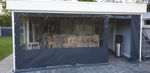Eine transparente Carport- oder Terrassenplane umschließt die Terrasse und schützt die Barhocker und Tische aus Holz. Ein großer Heizstrahler wärmt den gefliesten Boden. Rechts vervollständigen eine weiße Wand und eine kleine Treppe die Szene.