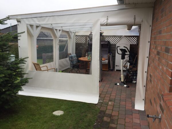Auf der Terrasse eines Backsteingebäudes steht ein weißer Carport oder eine Terrassenplane mit Fenster und Tür. Holzstühle, ein Tisch und Trainingsgeräte stehen auf dem Backsteinboden neben einem kleinen Schuppen und einem immergrünen Baum.