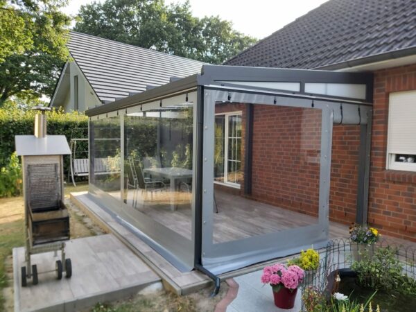 Ein Wintergartenanbau mit großen Glasflächen und einer Carport- oder Terrassenplane ist an ein Backsteinhaus angebaut. In der Nähe schmücken bunte Topfblumen den Bereich neben einem Außengrill und einem Teich, der von üppigem Grün umgeben ist.