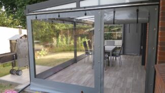 Eine moderne Außenterrasse verfügt über eine transparente, versenkbare Carport- oder Terrassenplane mit einem Fenster. Im Inneren befindet sich eine Essgruppe, während sich links außen ein Grill befindet. Topfblumen bereichern den Vordergrund.