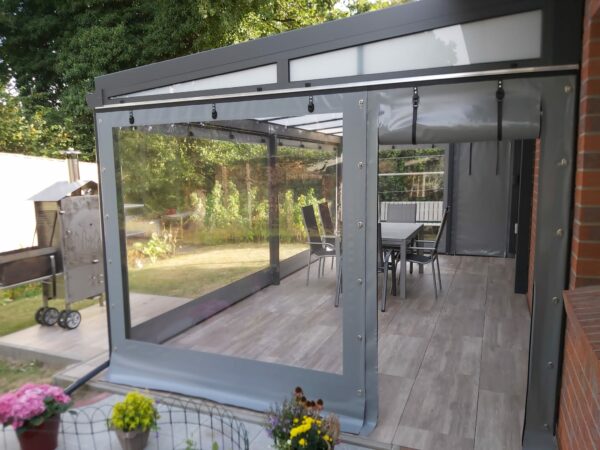 Eine moderne Außenterrasse verfügt über eine transparente, versenkbare Carport- oder Terrassenplane mit einem Fenster. Im Inneren befindet sich eine Essgruppe, während sich links außen ein Grill befindet. Topfblumen bereichern den Vordergrund.