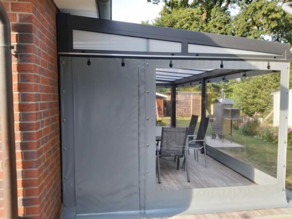 Eine überdachte Terrasse mit Carport- oder Terrassenplane und einem Fenster umschließt einen Essbereich mit Stühlen um einen Tisch mit Metallrahmen. Neben einer Backsteinmauer bietet er Ausblicke ins Grüne.