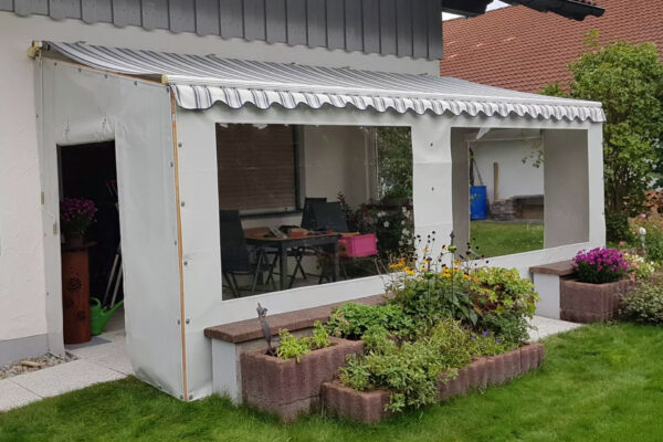 Ein weißer Hausanbau mit transparenten Carport- oder Terrassenplanen, überdacht von einer gestreiften Markise, blickt auf einen kleinen Garten. Im Inneren gibt es einen Tisch und Stühle inmitten von üppigem Gras und Grün.