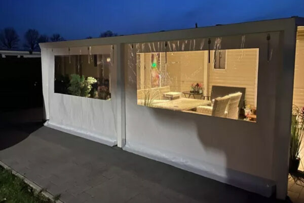Bei Einbruch der Dunkelheit erstrahlt eine gemütliche Terrasse mit Carport- oder Terrassenplane und durchsichtigen Kunststofffenstern in einem warmen Sitzbereich. Durch die transparenten Paneele ist der tiefblaue Himmel sichtbar.