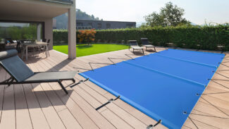 Eine moderne Außenterrasse mit Holzdeck verfügt über eine rechteckige Poolplane (Stangenabdeckung). Liegestühle stehen in der Nähe und ein Tisch steht überdacht. Grüne Hecken und ein entferntes Gebäude runden die Szene ab.