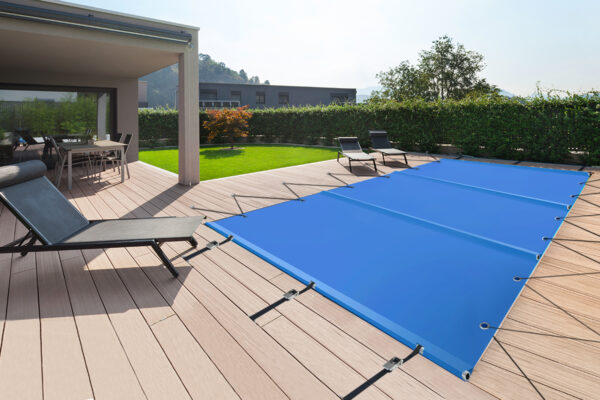 Eine moderne Außenterrasse mit Holzdeck verfügt über eine rechteckige Poolplane (Stangenabdeckung). Liegestühle stehen in der Nähe und ein Tisch steht überdacht. Grüne Hecken und ein entferntes Gebäude runden die Szene ab.