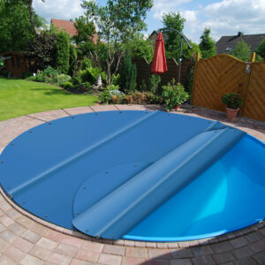 Ein Swimmingpool im Hinterhof, der teilweise mit einer blauen Plane abgedeckt ist. Der Pool ist von einer runden Ziegelterrasse, Grünflächen und einem Sichtschutzzaun aus Holz umgeben. Im Hintergrund sind ein roter Sonnenschirm und Gartenmöbel zu sehen.