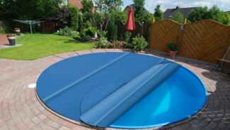 Ein Swimmingpool im Hinterhof, der teilweise mit einer blauen Plane abgedeckt ist. Der Pool ist von einer runden Ziegelterrasse, Grünflächen und einem Sichtschutzzaun aus Holz umgeben. Im Hintergrund sind ein roter Sonnenschirm und Gartenmöbel zu sehen.