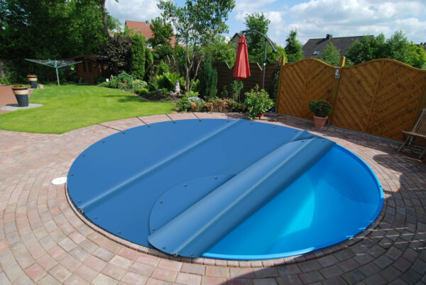 Ein Swimmingpool im Hinterhof, der teilweise mit einer blauen Plane abgedeckt ist. Der Pool ist von einer runden Ziegelterrasse, Grünflächen und einem Sichtschutzzaun aus Holz umgeben. Im Hintergrund sind ein roter Sonnenschirm und Gartenmöbel zu sehen.
