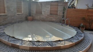 Ein kleiner, runder Pool, der mit einer runden Poolplanen-Stangenabdeckung abgedeckt ist, ist von Steinfliesen umgeben. Stufen führen hinauf, während eine Topfpalme und andere Pflanzen neben einem Holzzaun stehen.