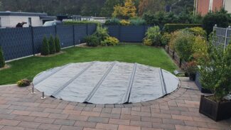 Eine runde Poolplanen-Stangenabdeckung bedeckt den runden Pool im Hinterhof, der von einer gepflasterten Terrasse, Rasen, Hecken und Sträuchern umgeben ist. Ein Holzzaun umschließt diese ruhige Gartenoase.