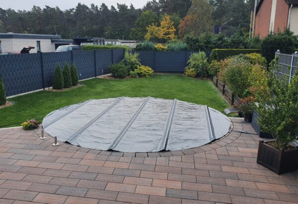 Eine runde Poolplanen-Stangenabdeckung bedeckt den runden Pool im Hinterhof, der von einer gepflasterten Terrasse, Rasen, Hecken und Sträuchern umgeben ist. Ein Holzzaun umschließt diese ruhige Gartenoase.