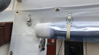 Eine Carport- oder Terrassenplane mit 2 Fenstern und 1 Tür ist neben einer Hauswand aufgerollt und gibt den Blick auf einen Teil eines Dekorationsgegenstands frei, der mit Riemen und Haken an einer weißen Struktur befestigt ist.