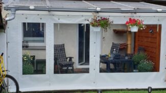 Ein kleiner weißer Wintergarten mit transparenten Paneelen verfügt über eine Carport- oder Terrassenplane mit zwei Fenstern und einer Tür. Im Inneren stehen karierte gepolsterte Stühle um einen Tisch mit Pflanzen. Blumenkörbe schmücken das Äußere, ergänzt durch einen gelben Schlauch draußen.