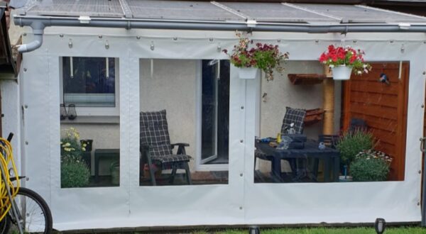 Ein kleiner weißer Wintergarten mit transparenten Paneelen verfügt über eine Carport- oder Terrassenplane mit zwei Fenstern und einer Tür. Im Inneren stehen karierte gepolsterte Stühle um einen Tisch mit Pflanzen. Blumenkörbe schmücken das Äußere, ergänzt durch einen gelben Schlauch draußen.