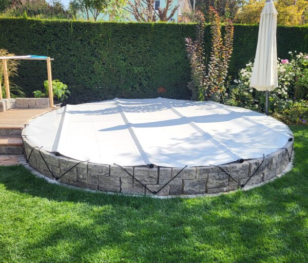 Ein runder Aufstellpool mit einer runden Poolplanen-Stangenabdeckung, die mit schwarzen Bändern befestigt ist. Umgeben von Gras, einer Steinmauer und üppigem Grün lugt rechts ein weißer Sonnenschirm hervor.