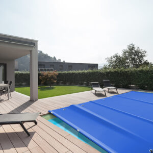Moderne Außenterrasse mit überdachtem Pool mit blauer Abdeckung. Auf dem Holzdeck stehen zwei schwarze Liegestühle und unter einer schattigen Pergola gibt es einen Essbereich. Eine grüne Hecke grenzt an den Platz und im Hintergrund sind Hügel zu sehen.