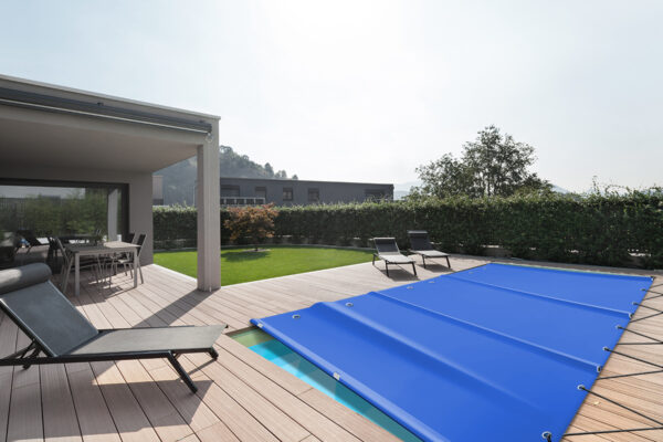 Moderne Außenterrasse mit überdachtem Pool mit blauer Abdeckung. Auf dem Holzdeck stehen zwei schwarze Liegestühle und unter einer schattigen Pergola gibt es einen Essbereich. Eine grüne Hecke grenzt an den Platz und im Hintergrund sind Hügel zu sehen.