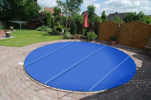 Eine runde Poolplanen-Stangenabdeckung deckt einen runden Hinterhofpool ab, der von Ziegelpflaster, üppigem Rasen, Pflanzen, einem Holzzaun und Gartenmöbeln umgeben ist.