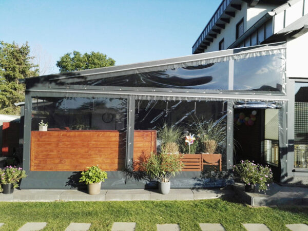 Eine moderne Terrasse mit einer transparenten „Terrassenplane in Dreiecksform mit Fenster (Links)“, Holzakzenten und Topfpflanzen, inmitten von Grün, im Hintergrund ist ein Teil eines Gebäudes sichtbar.