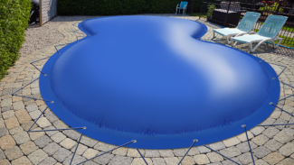 Auf einer Steinterrasse steht ein nierenförmiger Pool, der mit einer blauen, aufblasbaren Poolabdeckung abgedeckt und mit Seilen befestigt ist. Auf der rechten Seite stehen neben einer Hecke zwei weiße Liegestühle mit blauen Kissen.