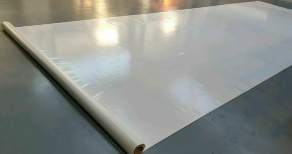 Eine große Rolle weißen, glänzenden Materials, teilweise ausgerollt auf einer glatten, reflektierenden Oberfläche. Der Glanz des Materials erinnert an eine Vinyl- oder Kunststoffzusammensetzung und das Sonnenlicht strömt sanft über die Oberfläche.