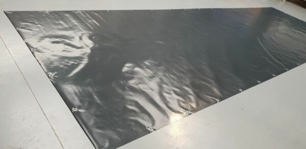 Eine große, glänzende, dunkelgraue Plane auf einem glatten, hellen Boden ähnelt LKW PVC Planen Reste. An den Rändern befinden sich Metallösen, die das Licht reflektieren und eine leicht faltige Textur aufweisen.