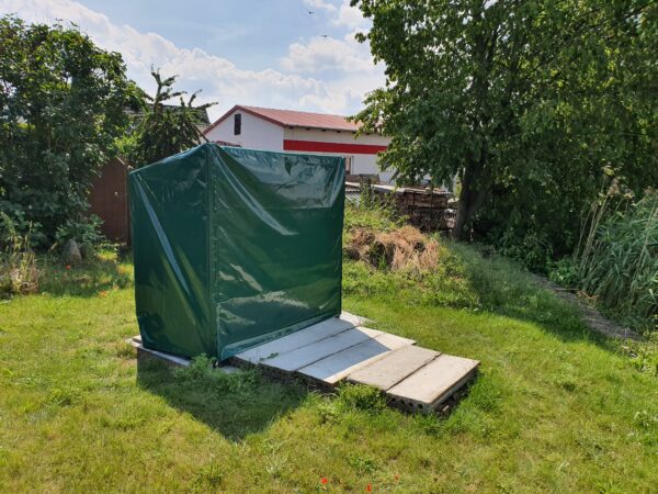Auf einer von Bäumen und Sträuchern umgebenen Rasenfläche steht ein begrünter Baukörper mit Schrägdach, ähnlich den Abdeckhauben Sonderform 1. Ein mit Betonplatten ausgelegter Weg führt dorthin, im Hintergrund ist ein Gebäude zu erkennen.