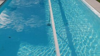 Ein langer, schmaler Pool mit klarem, blauem Wasser und einem Betondeck mit Leitern an einem Ende. Im Hintergrund ist eine Rasenfläche mit Liegestühlen zu sehen. Verschönern Sie ihn mit Schneeträger für Stangenabdeckungen!.