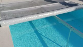 Ein Swimmingpool mit teilweise ausgefahrener silberner Abdeckung, zwei weißen Liegestühlen daneben und einem leuchtend orangefarbenen Schwimmkörper vor einem Zaun mit grünem Laub. Kaufen Sie noch heute Schneeträger für Stangenabdeckungen!.