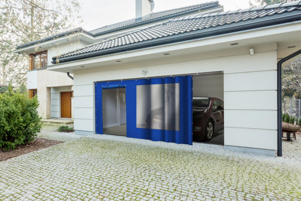 Modernes Haus mit sichtbarer Garage mit Schiebeplane mit Fenster/mit Tür. Im Inneren ist die Vorderseite eines Autos geparkt. Die Außenseite hat ein Ziegeldach, eine gepflasterte Auffahrt und Büsche auf der linken Seite.