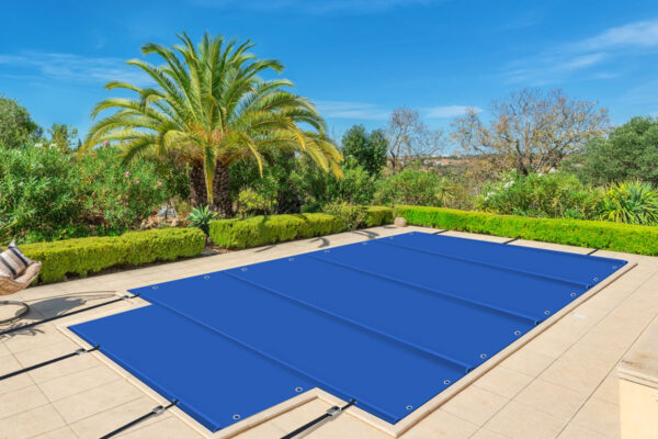 Eine rechteckige Poolplane + Treppe (Stangenabdeckung) ist auf einem Pool in einem sonnigen Hinterhof mit gefliester Terrasse befestigt, umgeben von gut geschnittenen Hecken, üppigen Pflanzen und einer großen Palme unter einem klaren blauen Himmel.