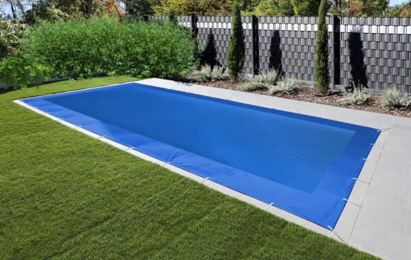 Ein rechteckiger Swimmingpool mit rechteckiger Netzabdeckung verfügt über eine Steinterrasse und üppiges grünes Gras. Hohe Pflanzen und ein Sichtschutzzaun rahmen den Hintergrund unter einem klaren Himmel ein.